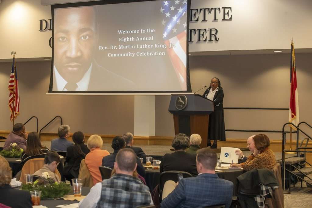 person at a podium addressing an audience with a screen with text that reads"Welcome to the Eighth Annual Rev. Dr. Martin Luther King Jr. Community Celebration
